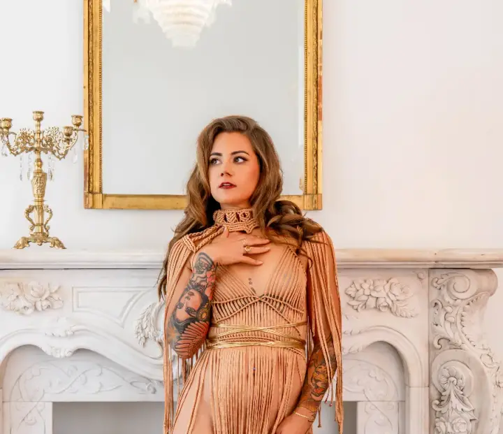 Helena Herndon in a gold gown, hand on her chest, in front of a marble fireplace