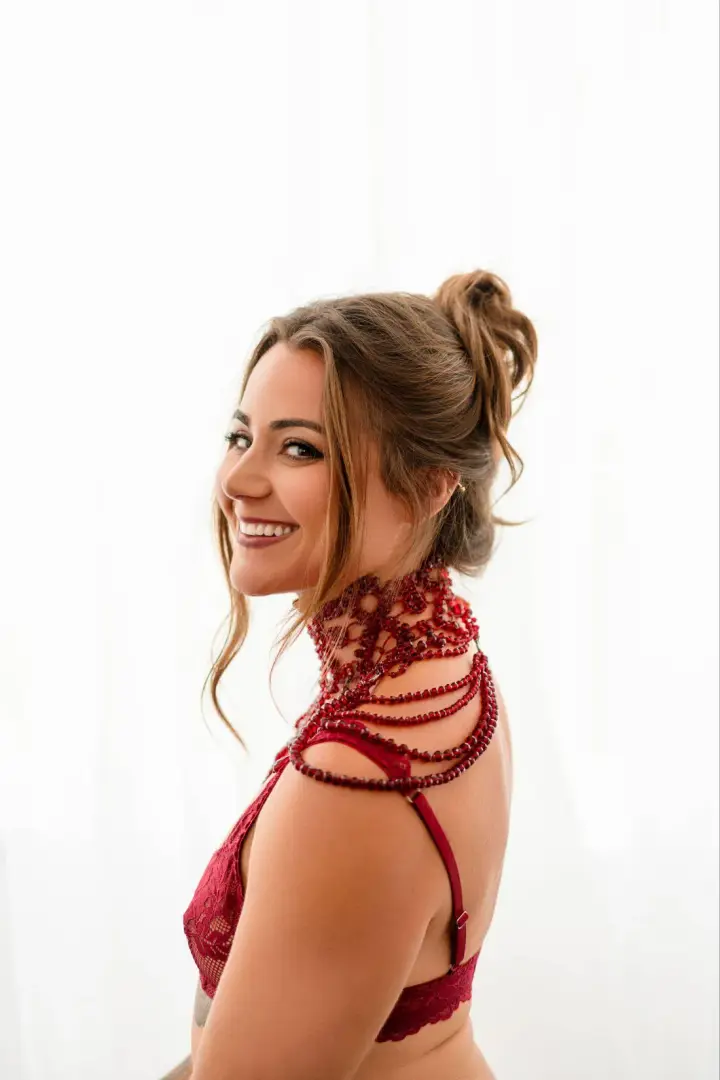 Helena Herndon in red lace lingerie and a beaded red collar, smiling over her shoulder